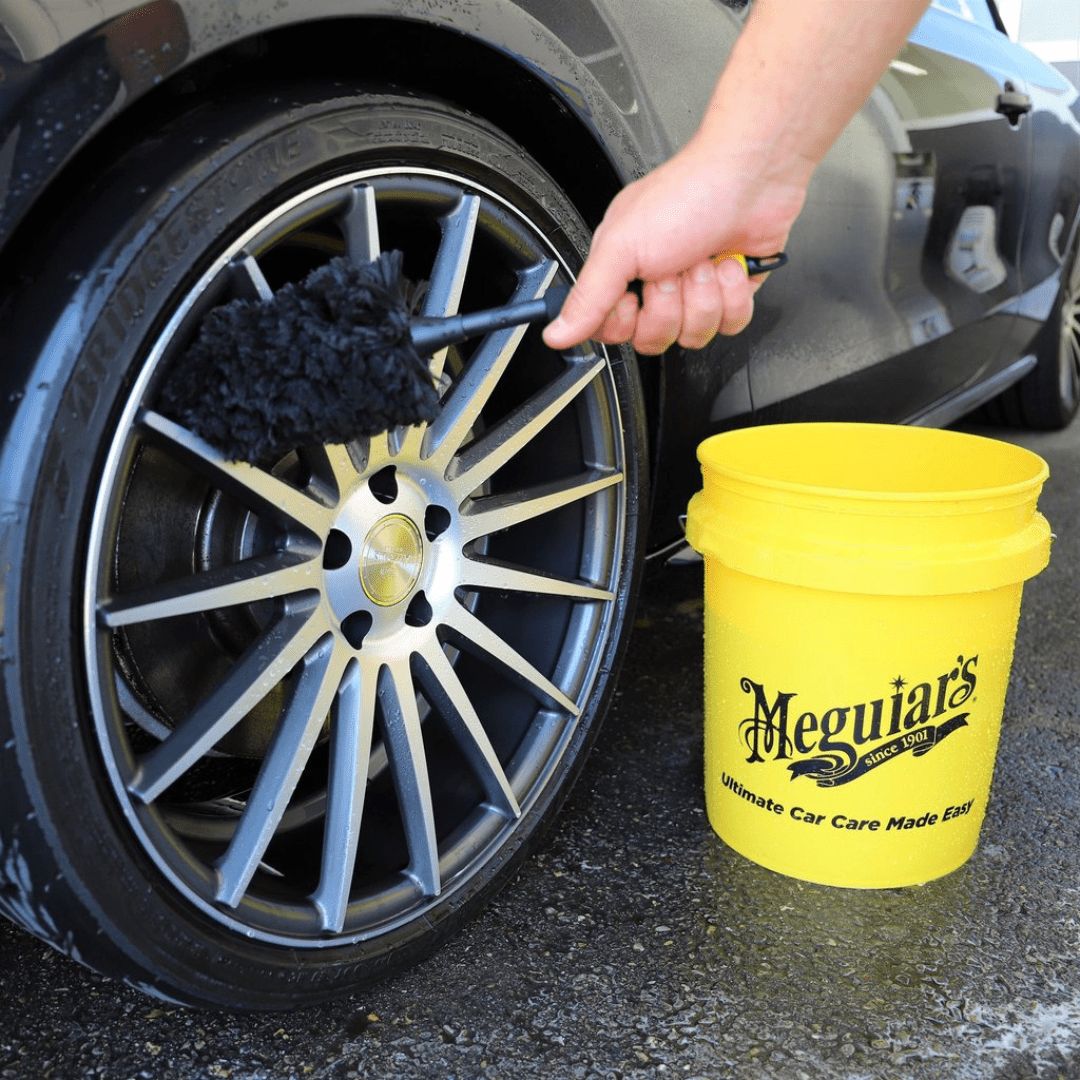Die Meguiars Supreme Wheel Brush Medium Felgenbürste zwischen den Speichen einer Felge mit Wascheimer daneben