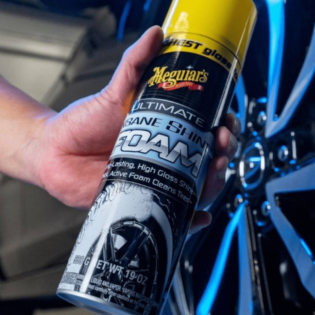 Die Meguiars Ultimate Insane Shine Foam Reifenpflege Sprühdose in der Hand vor einem Autoreifen